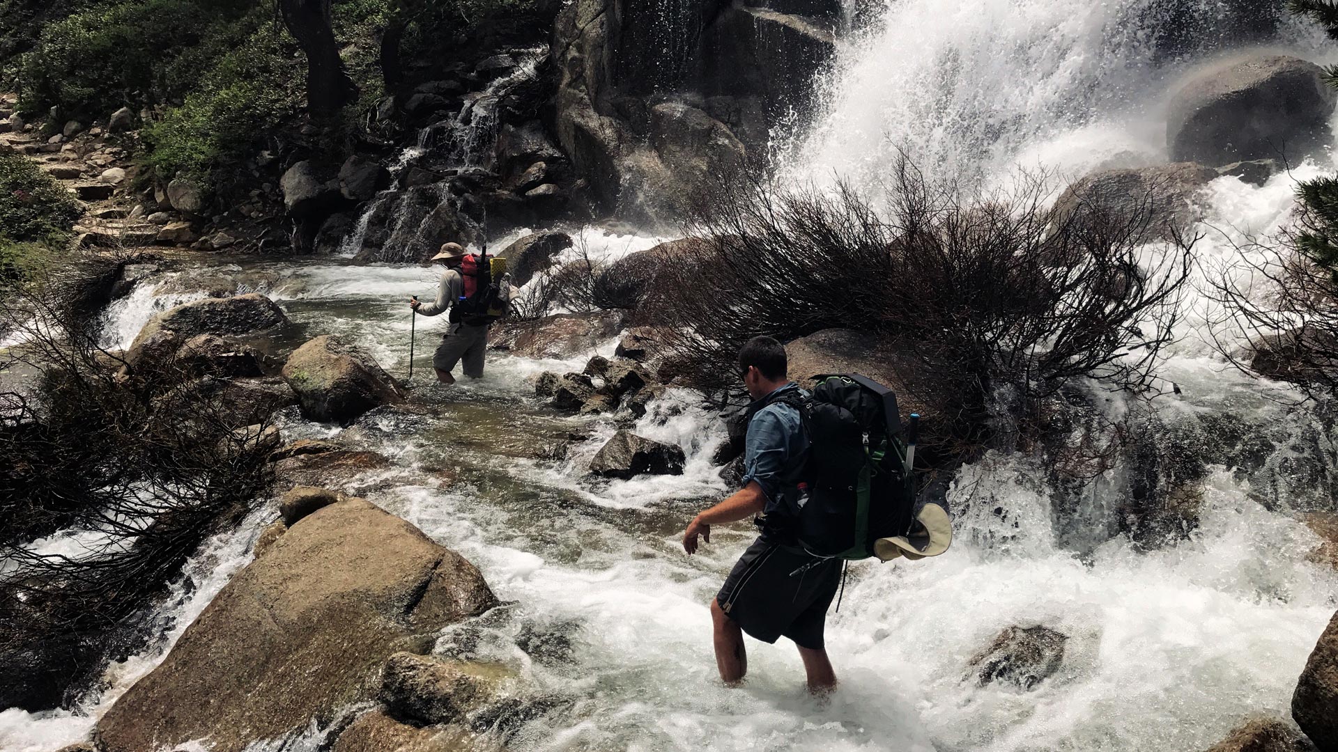 Two hikers attempt a safe crossing of a river while backpacking.