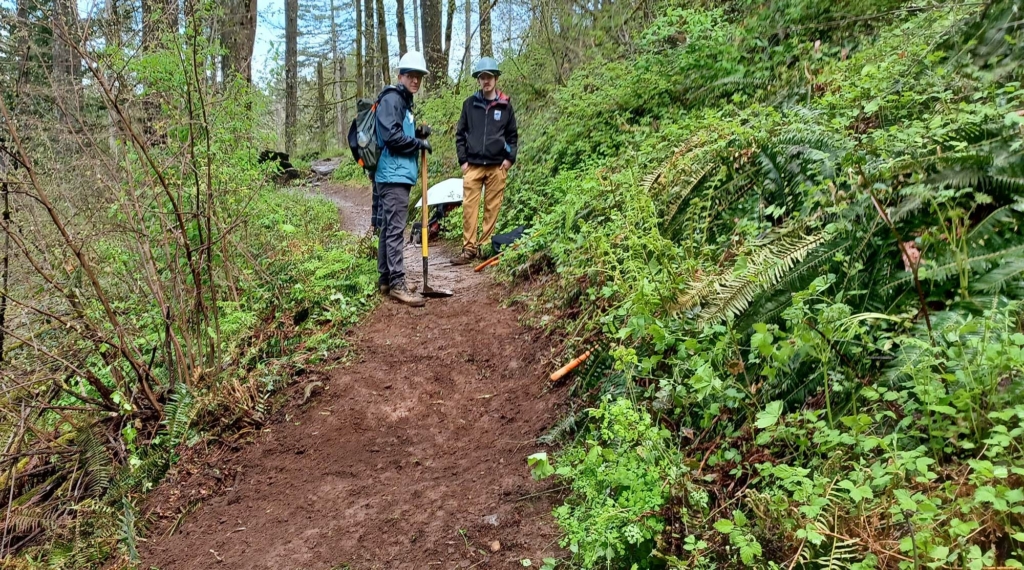 Two people stand on a dirt trail in the forest, one holds a trail tool