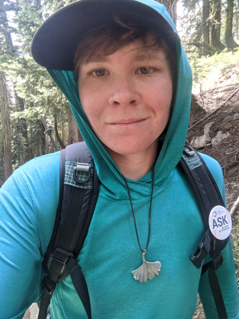 A portrait of a hiker on the trail with a pin reading "Ask me about my pronouns"